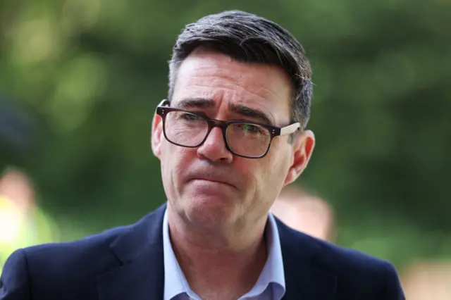 Andy Burnham wears a dark suit and light coloured shirt. He has glasses and dark brown hair. His lips are tightly together.