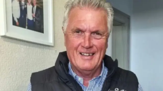 David Moss stands in a room while wearing a blue stripped shirt and black gilet