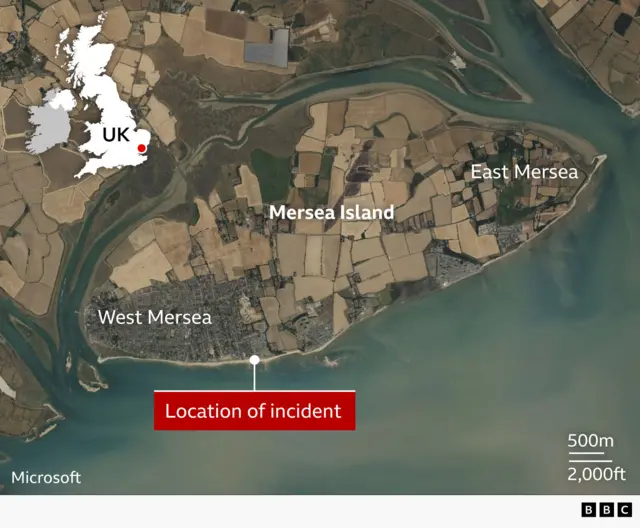 Satellite map of Mersea Island in Essex. The towns of West Mersea and East Mersea are labelled at either end of the island and a red marker shows the location of the incident on the island's southern shoreline at the eastern edge of West Mersea. An inset map of the UK shows Mersea Island's position in southeast England.