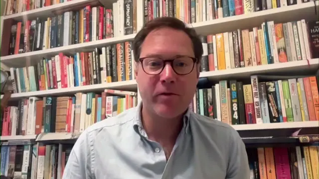 Allen Simpson, a man, wearing a shirt with a bookshelf behind him.