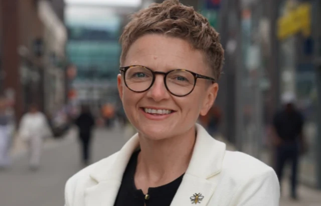 Bev Craig has short blonde hair and wears dark-framed round glasses. She wears a white jacket over a black top