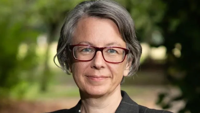 Geraldine Coggins has grey, parted hair and wears purple framed glasses and a dark jacket.