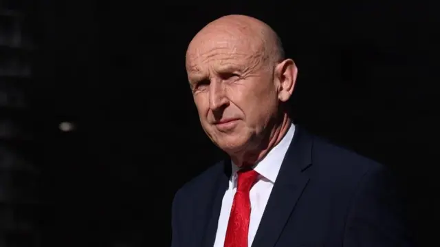 John Healey wears a dark-coloured suit, white shirt and a red tie