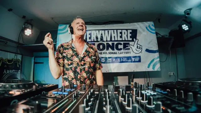 Fatboy Slim wears a floral shirt while standing behind a set of DJ decks.