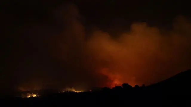 Flames and smoke are illuminated on a distant hillside against a black sky.