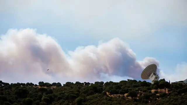 Smoke from a nearby fire drifts across the horizon of a hill with dense trees and a Nasa antenna