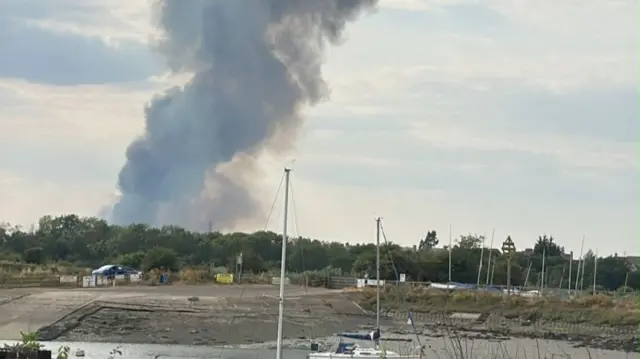 Huge plumes of smoke rising above a line of trees, which are by a riverbed with boats on it.