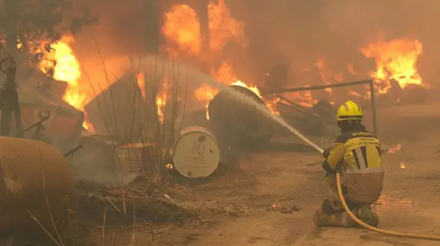 A firefighter is shown crouched on the ground, spraying water into flames and thick smoke enveloping barrels and tyres.