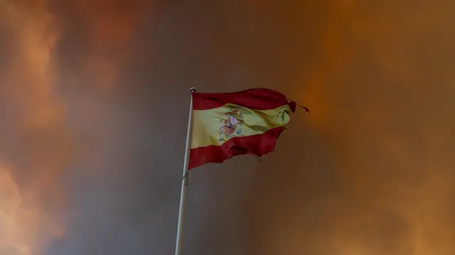 A Spanish flag (three horizontal stripes: red, yellow, and red, with the yellow stripe twice as wide as each red stripe. It also includes the national coat of arms shifted slightly toward the hoist side) partially burned with dark smoke clouding the sky