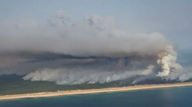 An aerial view shows a large wildfire spreading a number of miles across a coastal area behind a beach.