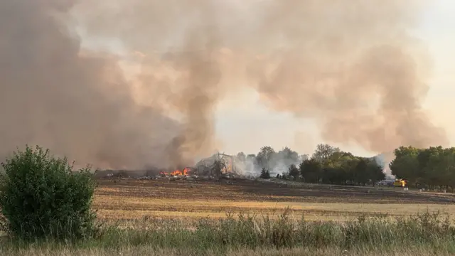 Fire burning around a collapsed building on a field, pictured further away than the other picture.