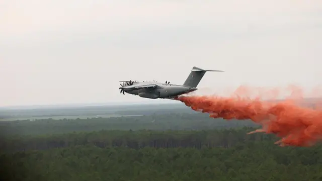 Red fire retardant is dropped out the back of an Airbus A400M. A red trail has formed behind the plane while green trees are seen below