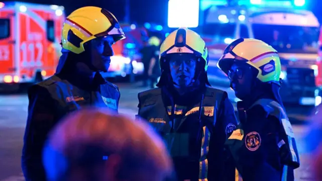 Several first responders are seen talking with emergency vehicles seen behind them