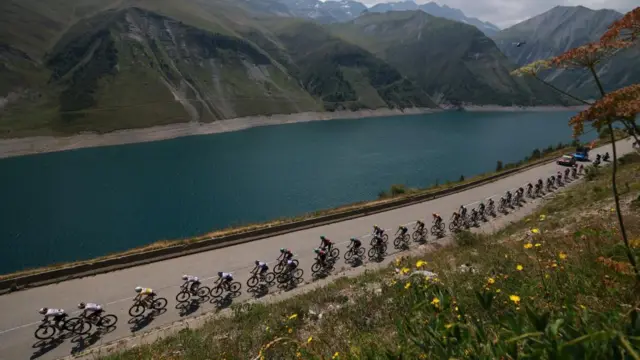 Cyclists move along a road next to a lake during the 20th stage of the Tour De France. The foot hills and mountains of the Alps can be seen in the background