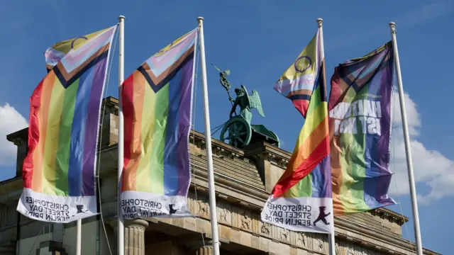 Pride flags move in the wind in front of the Brandenburg Gate in Berlin, the sky is blue with a few clouds.