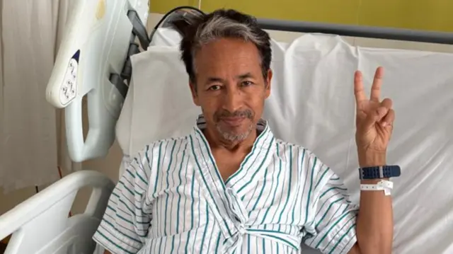 Soman Wangchuk lies on a hospital bed making a victory symbol