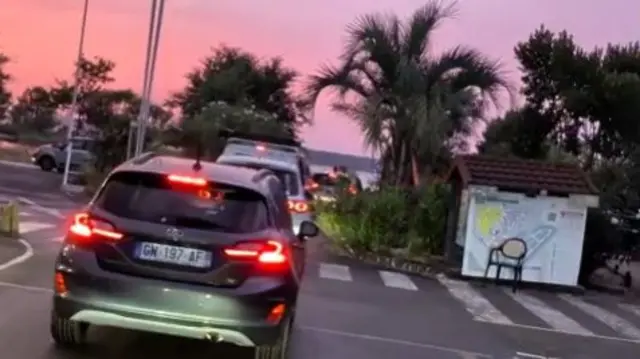 A queue of cars with a pink sky that is glowing from flames
