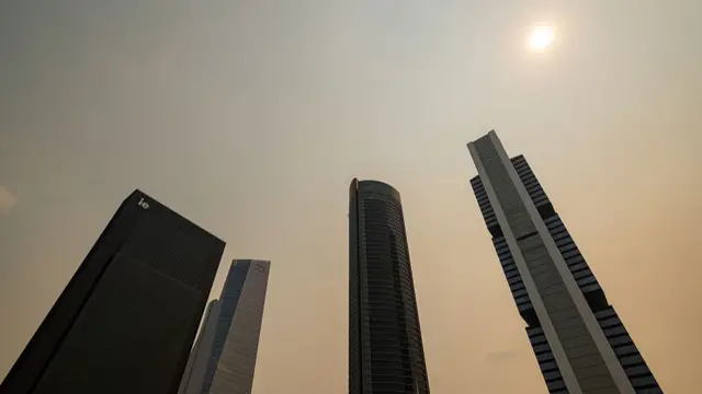 A cluster of skyscrapers are shown from ground level, with the sun visible but partially obscured through smoke.