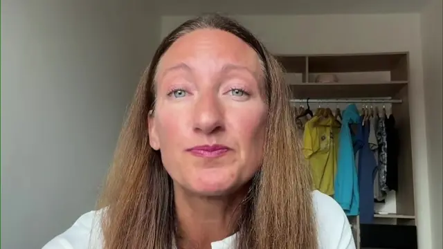 Rebecca Papin speaks into a web camera. She is wearing a white top and a clothes rack with clothes can be seen behind her