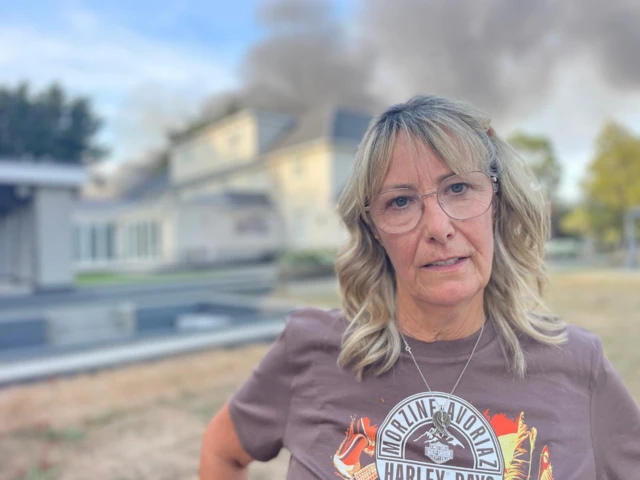 A woman with blonde hair and glasses looks into the camera. She is standing in front of a large house and pool. Behind the house large plumes of grey smoke are going into the air.
