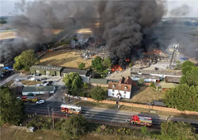 Aerial shot shows huge plumes of black smoke inthe air. Fire engines are parked on the closest road. Orange flames are visible through the smoke.