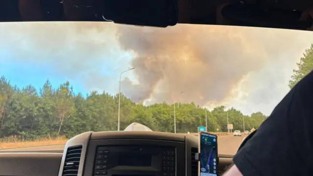 Smoke rises above a tree line next to a motorway in France. The picture has been taken from the inside of a car and shows a driver's arm and a phone with a maps app displayed on it