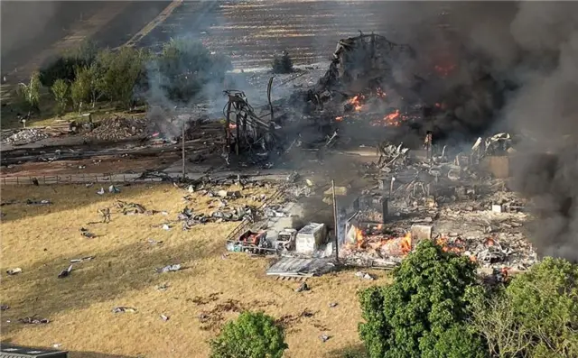 Pockets of orange flames can be seen in the wreckage of a collapsed and burnt out building. Thick black smoke is rising from the debris.