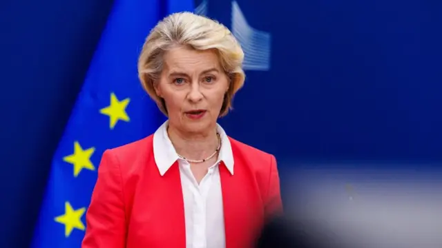 European Commission President Ursula von der Leyen wears a white coloured shirt and a pink jacket. She is standing in front of an EU flag