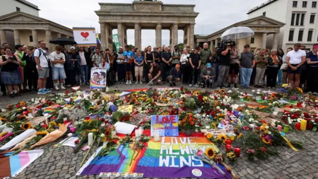 A large group of people is shown gathered at Berlin's Brandenburg gate behind Pride flags and bunches of flowers on the ground.