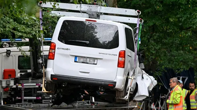 A white van is shown being lifted onto the back of a flatbed truck, as several men in hi-viz jackets watch from the side.
