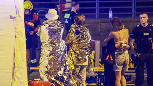 Two people are wrapped in silver security blankets. Another person hugs someone nearby as a police officer stands nearby