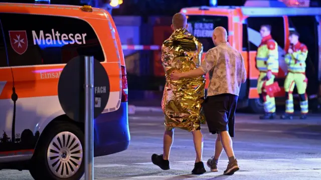 A man wrapped in a silver blanket walks away from the camera. A man next to him has an arm around him