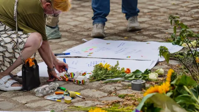 A woman writes a message on white card. There are a range of rainbow coloured pride stickers next to it and a number of sunflowers nearby