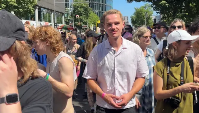 Nico Nölken stands in the middle of the crowd at Berlin Pride. He is wearing a striped white shirt and has short blonde hair and a moustache