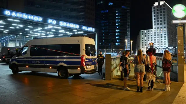 A group of people stand next to a staircase to a station in Potsdamer Platz in Berlin. A police van can be seen behind some tape to their left