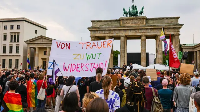 Hundreds of people are shown gathered in front of the Brandenburg Gate, with Pride flags and protest signs. One reads "von trauer zu wut zu widerstand", which translates as "from grief to anger to resistance"