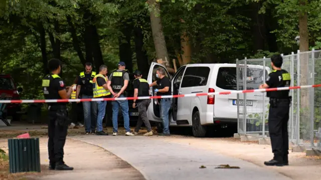 A man wearing blue gloves and a surgical mask is seen next to the open door of a white van. Several uniformed police officers stand near the vehicle. The path the vehicle is on has been cordoned off and two officers stand guard near some police tape