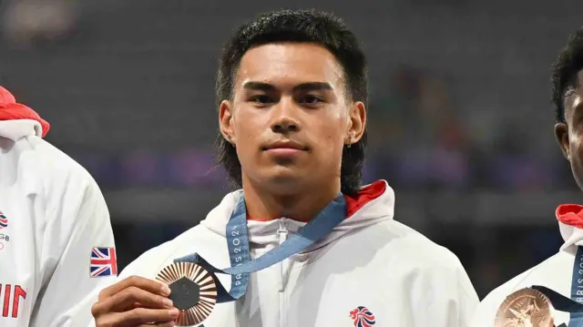 Louis Hinchliffe, wearing Team GB's white 2024 Olympics jacket, holds up his bronze medal in Paris