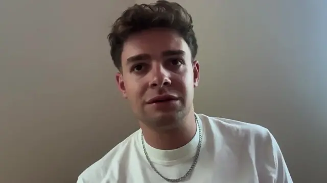 Julian Miethig wears a white t-shirt and a silver chain as he speaks to a webcam. He is against a white background
