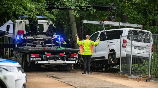 A van being hoisted up as a flat bed truck waits