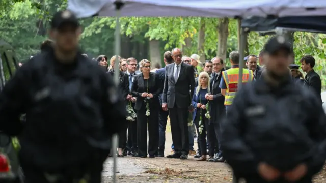 Merz standing in a group of people who wear black, holding white roses by their sides. Police stand in front of them