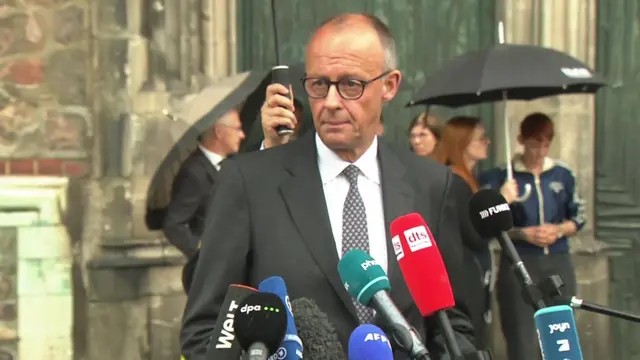 Friedrich Merz is showing speaking behind a bank of microphones, wearing a suit and tie