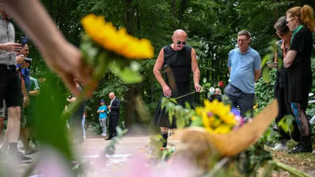 A man in a black t-shirt and a black kilt lays a rose near tributes in Tiergarten park. Several people can be seen holding sunflowers