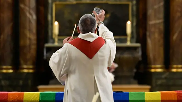A priest is shown with his back to the camera, lighting a candle with a rainbow flag over the table in front of him.