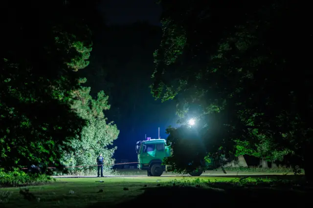 A police truck illuminates Tiergarten park at night as an officer watches on.