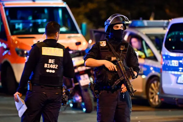 A police officer holding an automatic rifle and wearing a helmet is pictured in front of several police vehicles, with another office beside him with his back to the camera.