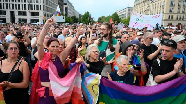 A crowd holding Pride flags looking downcast