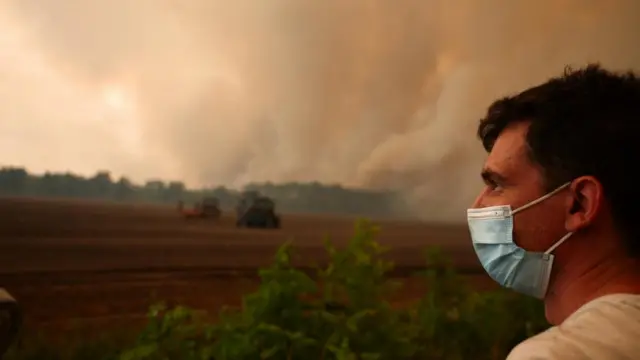 A man wears a mask, while smoke rises from a wildfire in Gironde