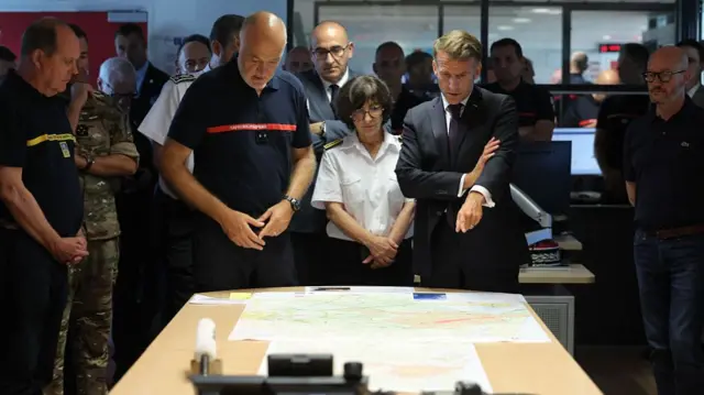 Emmanuel Macron looks at maps on a table, flanked by firefighting officials. He is pointing with his left hand.