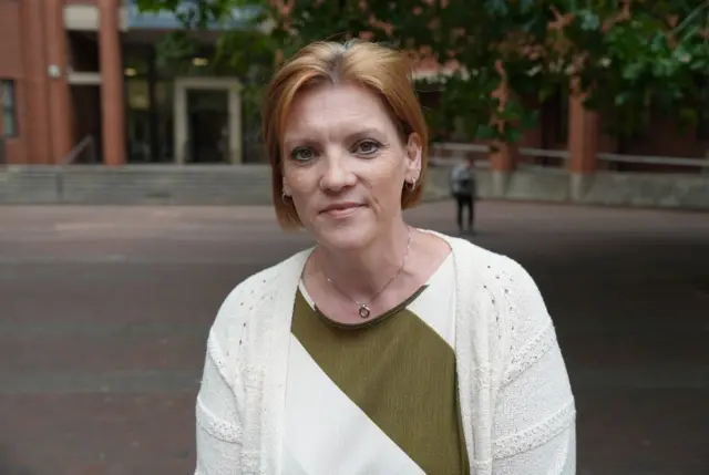 A woman with a blonde bob is looking down to the side and wearing a white cardigan, green and white top and silver necklace. The background is blurred.